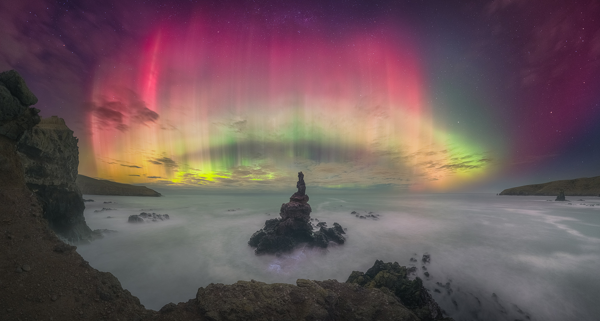 A bright aurora in purple, yellow, and green is visible in the sky as waves are seen moving in fron of cliffs.